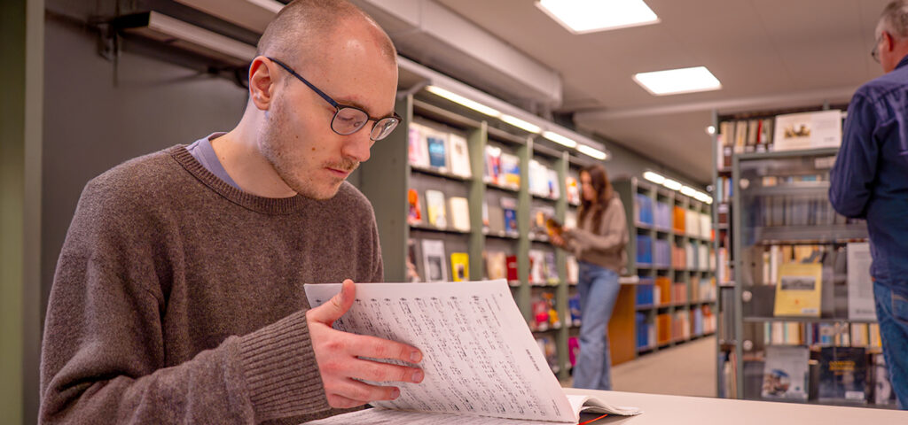 Ung man bläddrar i noter på Musik- och teaterbiblioteket. Två personer rör sig i bakgrunden.