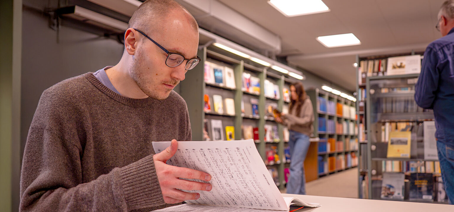 Ung man bläddrar i noter på Musik- och teaterbiblioteket. Två personer rör sig i bakgrunden.