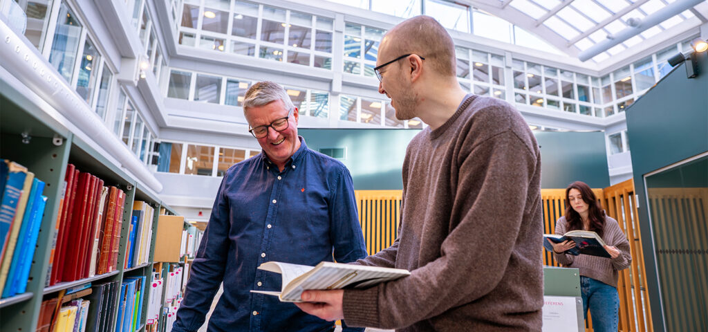 Två personer står vid en bokhylla i ett ljust bibliotek och bläddrar i böcker. I bakgrunden står ytterligare en person och läser.