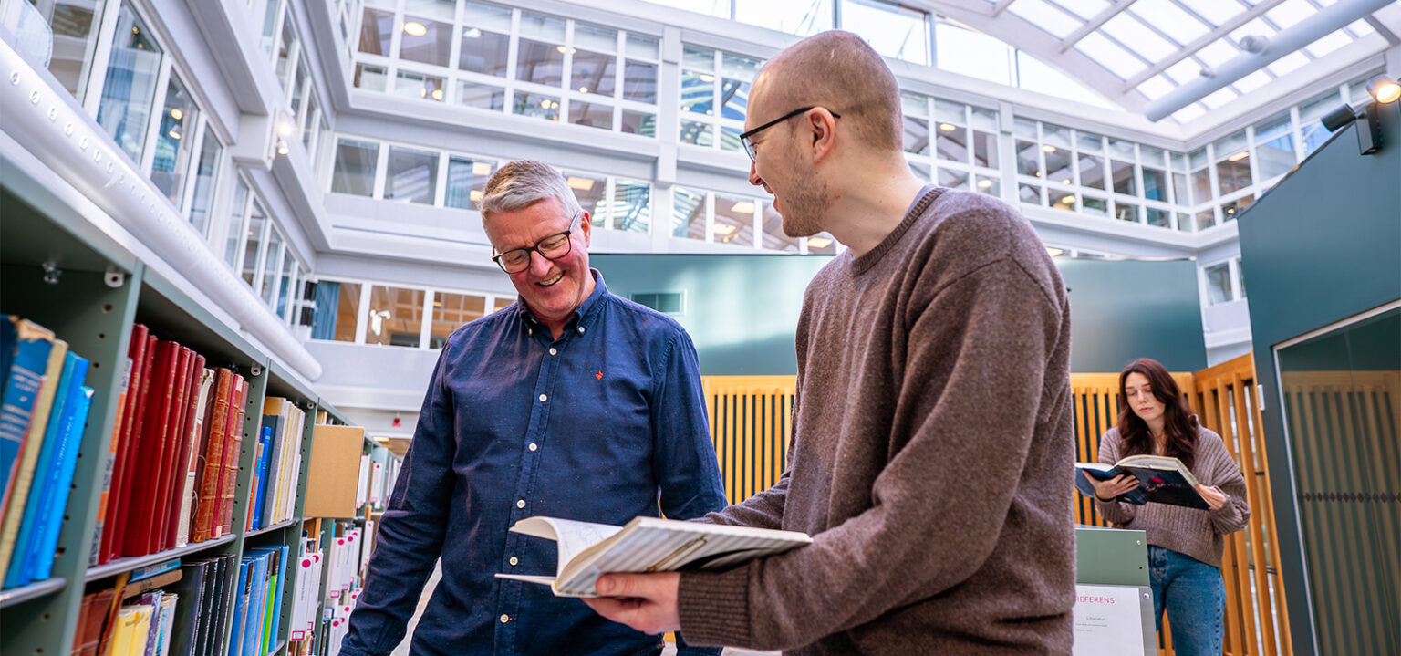 Tv&aring; personer st&aring;r vid en bokhylla i ett ljust bibliotek och bl&auml;ddrar i b&ouml;cker. I bakgrunden st&aring;r ytterligare en person och l&auml;ser.